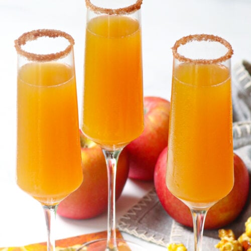Three champagne flutes filled with golden apple cider mimosas, cinnamon sugar-rimmed glasses, fresh apples in background for Apple Cider Mim