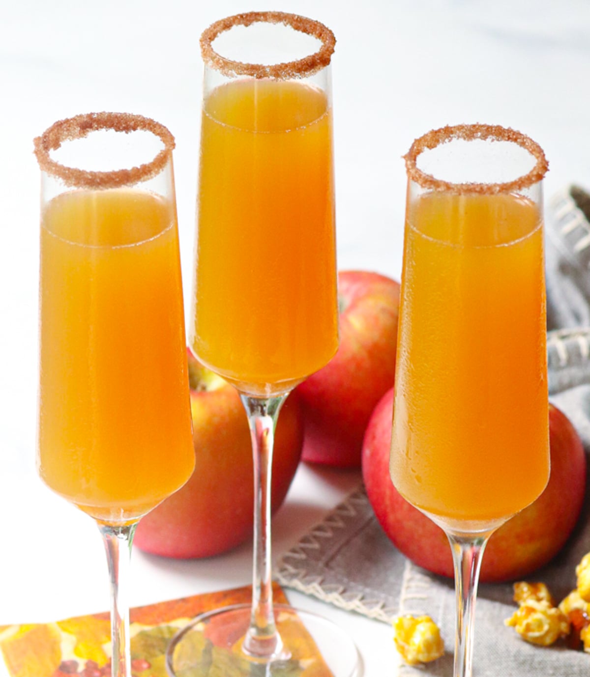 Three champagne flutes filled with golden apple cider mimosas, cinnamon sugar-rimmed glasses, fresh apples in background for Apple Cider Mim