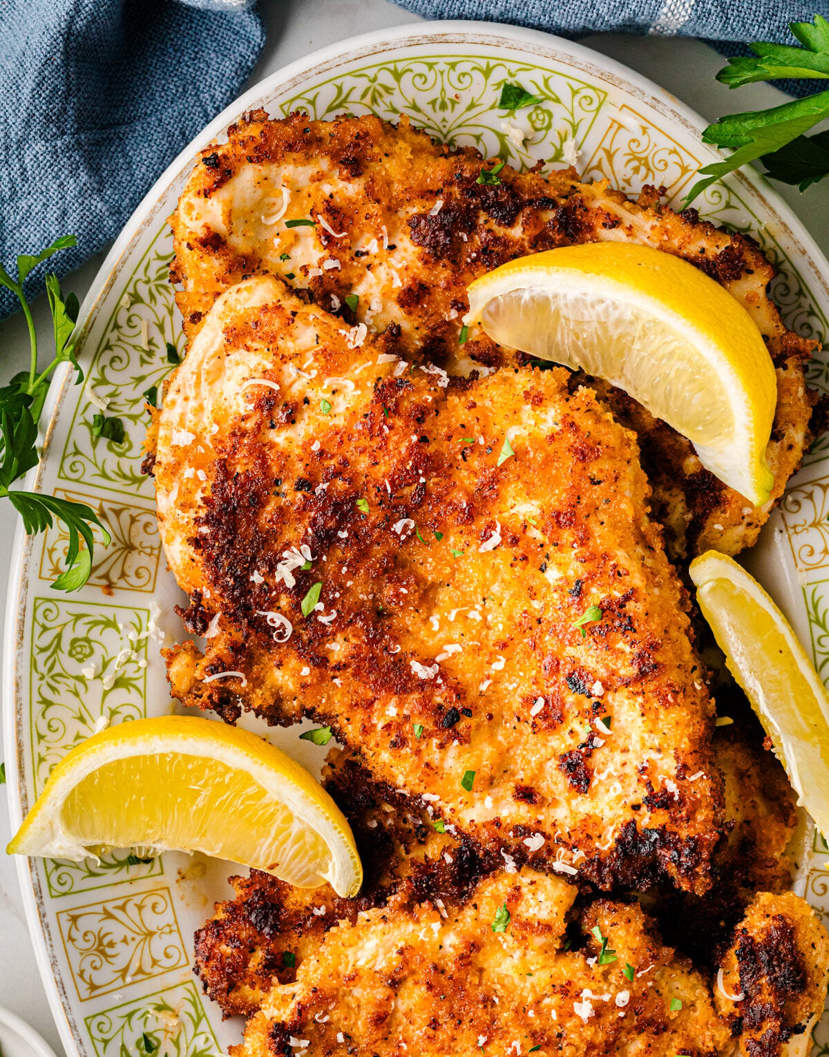 Parmesan crusted chicken breasts on decorative plate with lemon wedges and fresh parsley garnish, overhead view