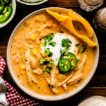 Overhead view of white chicken chili 9 in a speckled bowl, topped with sour cream, jalapeños, cilantro, and tortilla chips, surrounded by li