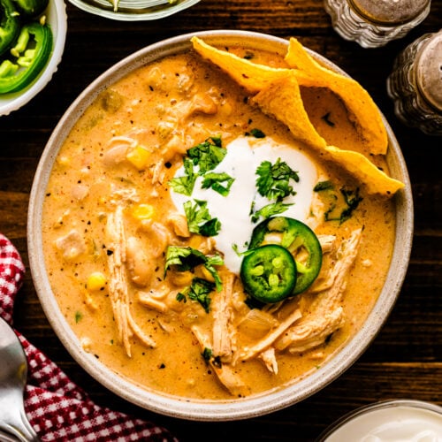 Overhead view of white chicken chili 9 in a speckled bowl, topped with sour cream, jalapeños, cilantro, and tortilla chips, surrounded by li