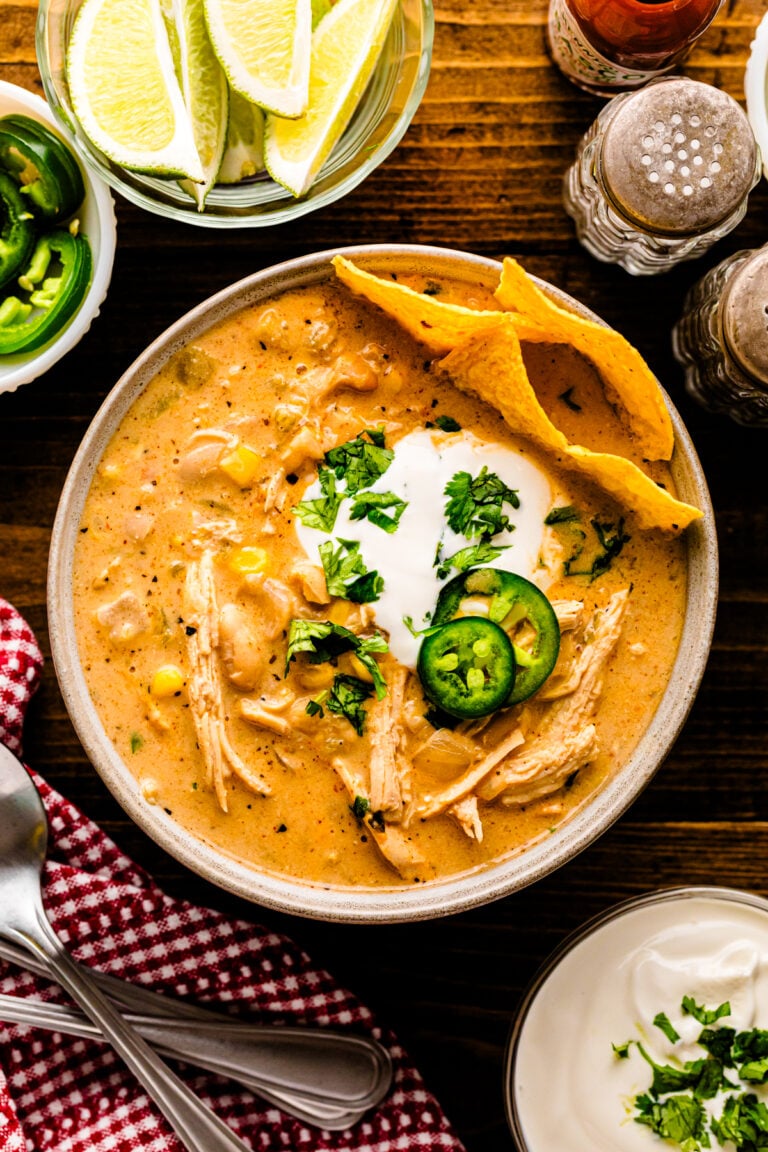 Overhead view of white chicken chili 9 in a speckled bowl, topped with sour cream, jalapeños, cilantro, and tortilla chips, surrounded by li