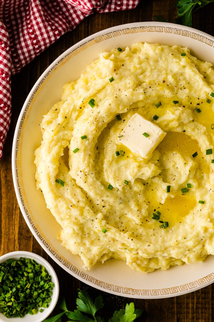 Overhead shot of a serving bowl filled with creamy, swirled Boursin mashed potatoes, melted butter pooling in the center and fresh chives scattered on top.
