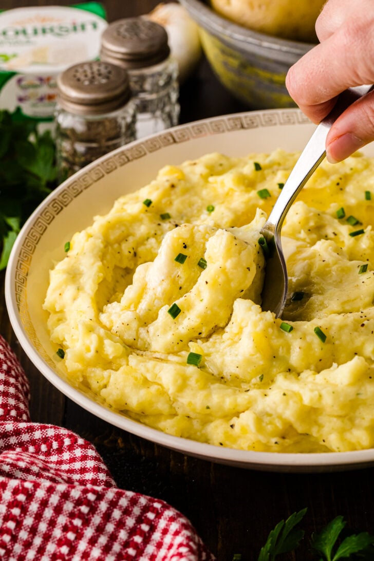 Close-up of extra-creamy mashed Yukon Gold potatoes mixed with Boursin garlic-herb cheese, topped with melted butter and chives as a spoon lifts a serving.