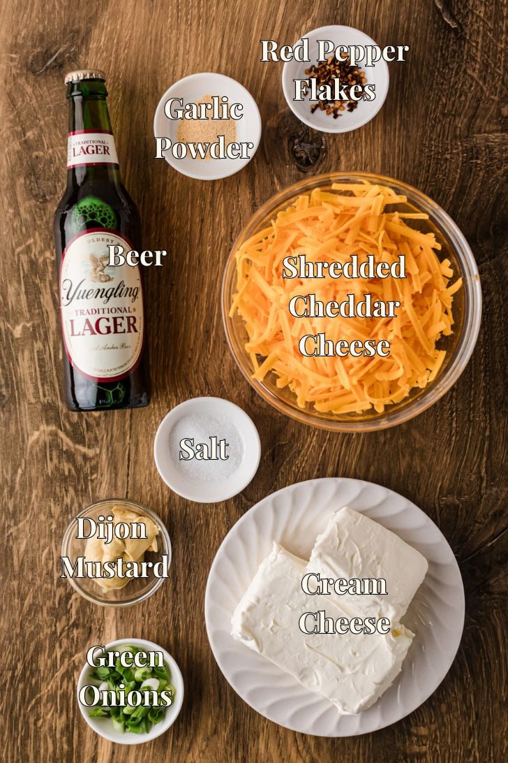 Ingredients for Crock Pot beer cheese dip on the counter—cream cheese, shredded sharp cheddar, lager beer, Dijon mustard, garlic powder, red pepper flakes, salt, and green onion.