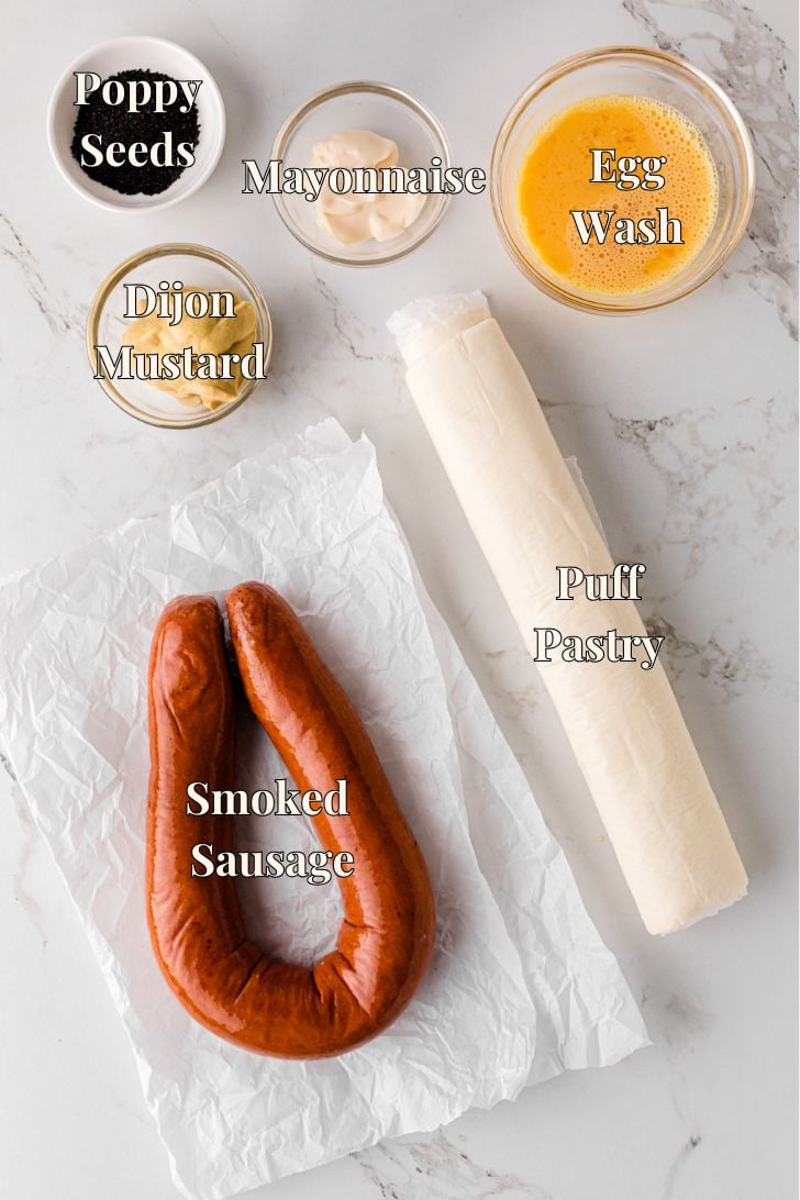 Ingredients for big pigs in a blanket on a counter, including puff pastry, smoked sausage, Dijon mustard, mayonnaise, egg, and poppy seeds.