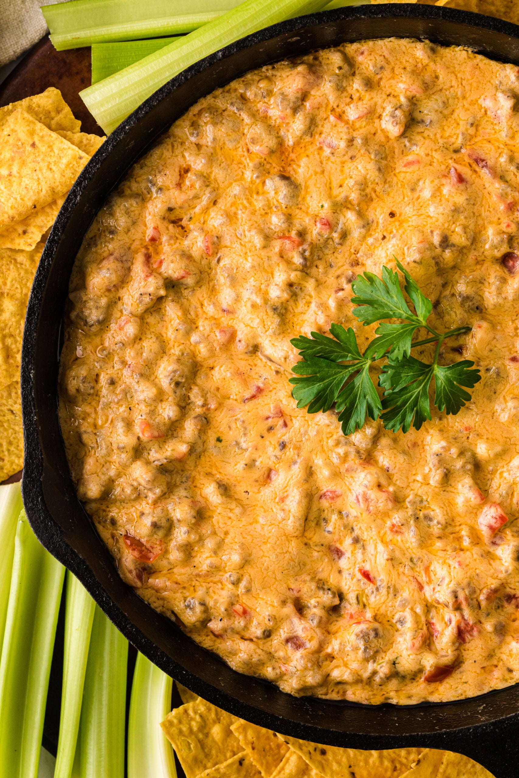 Pimento Cheese and Sausage Dip 5 in black cast iron skillet with tortilla chips and celery sticks on the side, garnished with fresh parsley