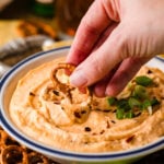 Hand dipping pretzel into beer cheese dip garnished with green onions in white bowl with blue rim