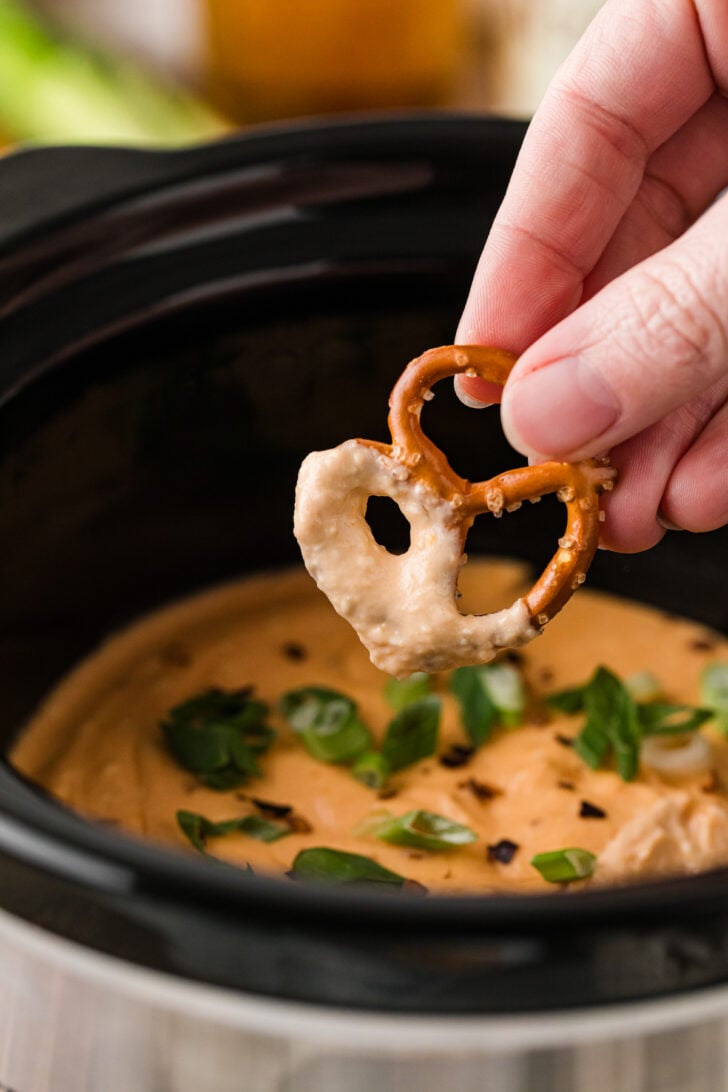 Hand dipping a pretzel bite into warm Crock Pot beer cheese dip, showing the thick, creamy texture as it coats the pretzel.
