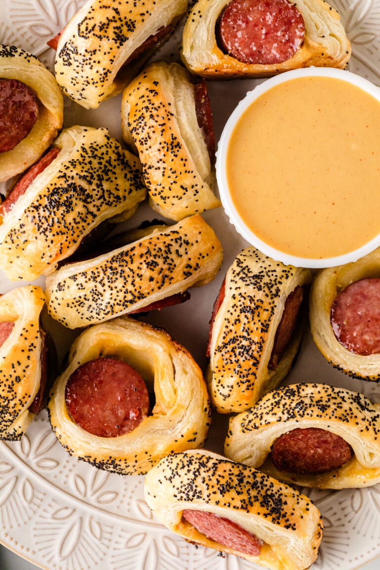 Big Pigs In A Blanket (Puff Pastry + Smoked Sausage) with poppy seeds arranged on a plate with cheese dip
