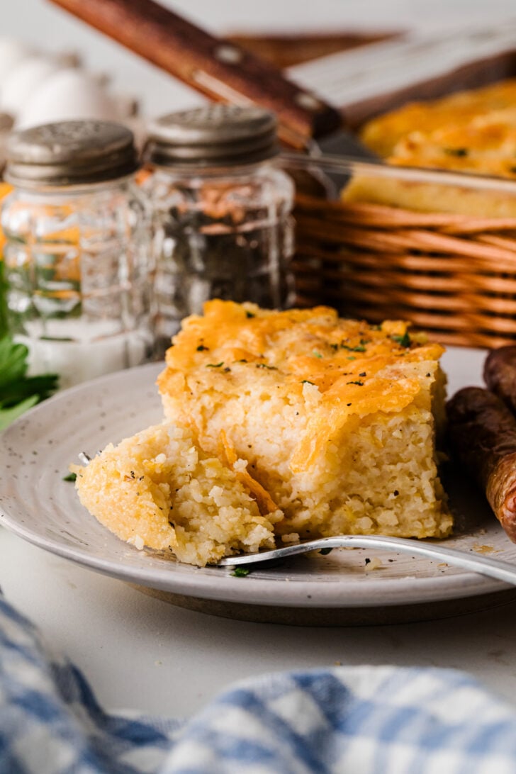 Square of baked cheese grits on a white plate with sausage links, easy Southern breakfast with a make‑ahead cheese grits casserole.