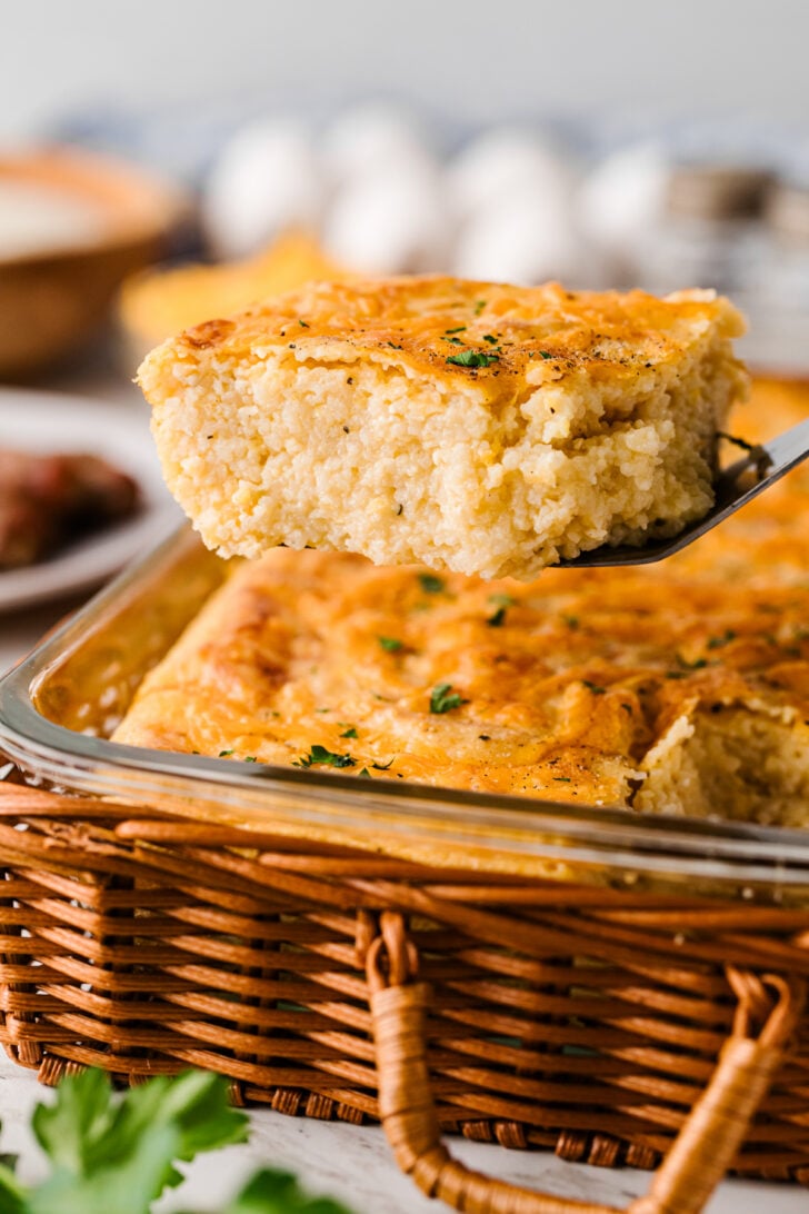 Square of baked cheese grits lifted on a metal spatula, showing creamy inside and crispy golden edges of this Southern casserole.