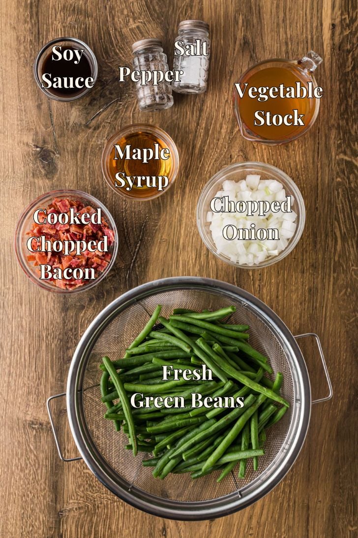 Ingredients for crock pot maple bacon green beans laid out on a counter, including green beans, bacon, maple syrup, broth, and seasonings.