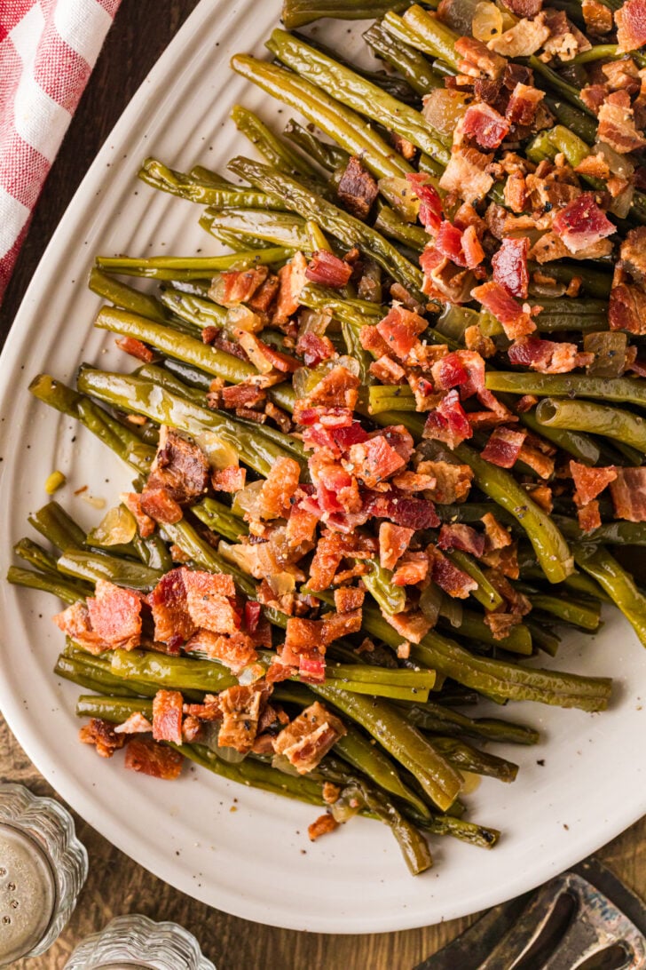 Slow‑cooked maple bacon green beans piled on a white serving platter with crisp bacon on top.