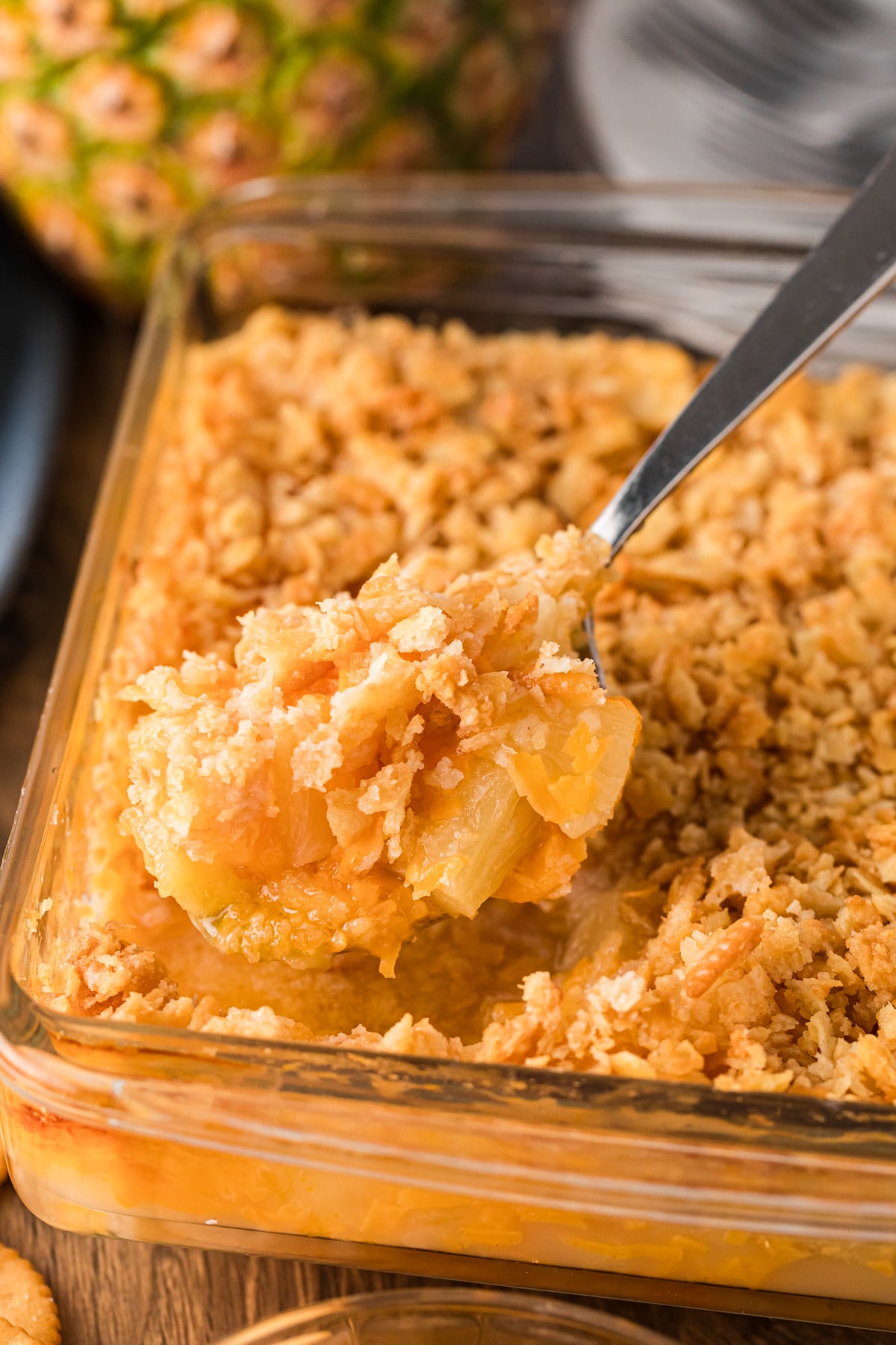 Spoon lifting golden pineapple casserole with crushed cracker topping from glass baking dish