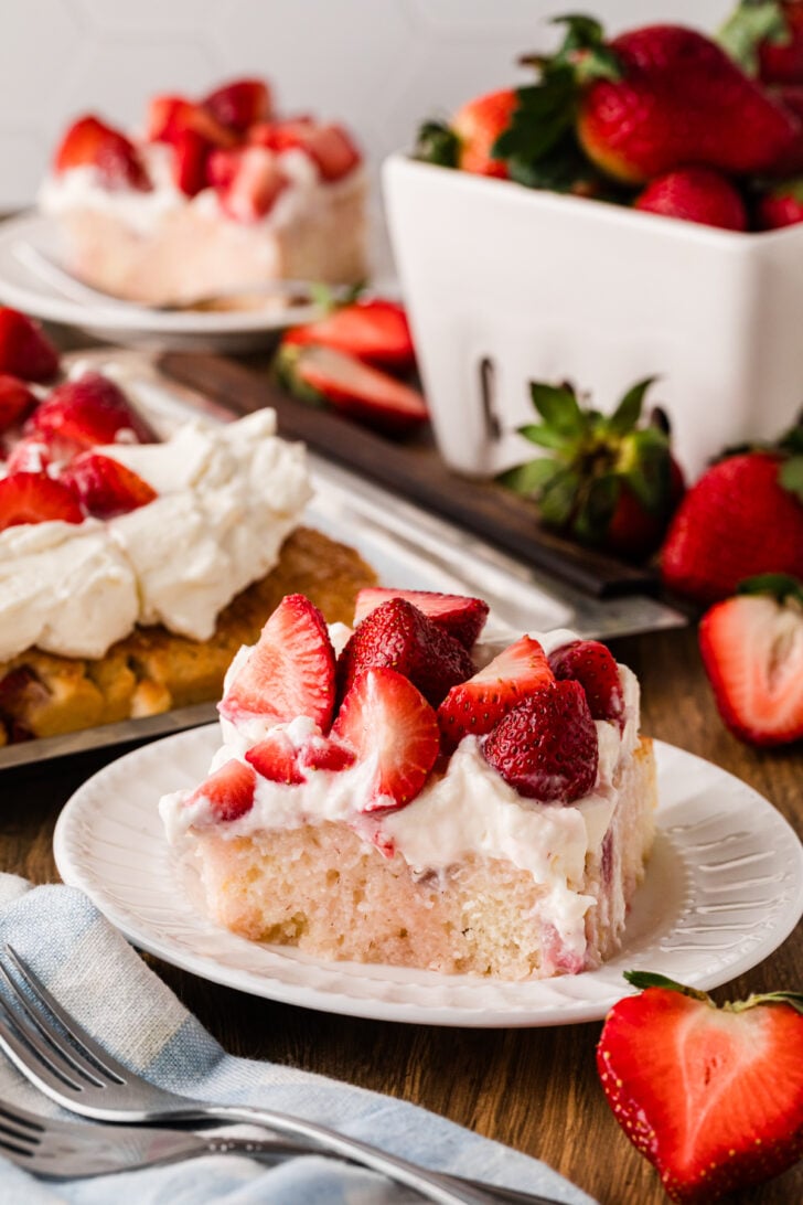 Strawberry Shortcake Sheet Cake slice on white plate with whipped cream and fresh strawberries, fork beside, whole cake and berries in backg