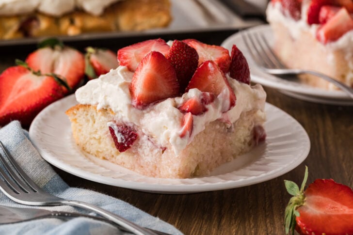 Strawberry Shortcake Sheet Cake slice on white plate with whipped cream, fresh strawberries, fork, and napkin on dark wood table