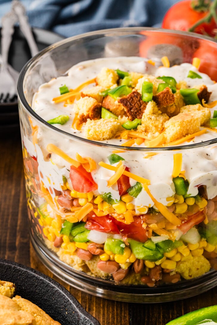 Southern Cornbread Salad layered in a glass bowl with cornbread crumbles, beans, corn, peppers, tomatoes, cheese, and creamy dressing topped
