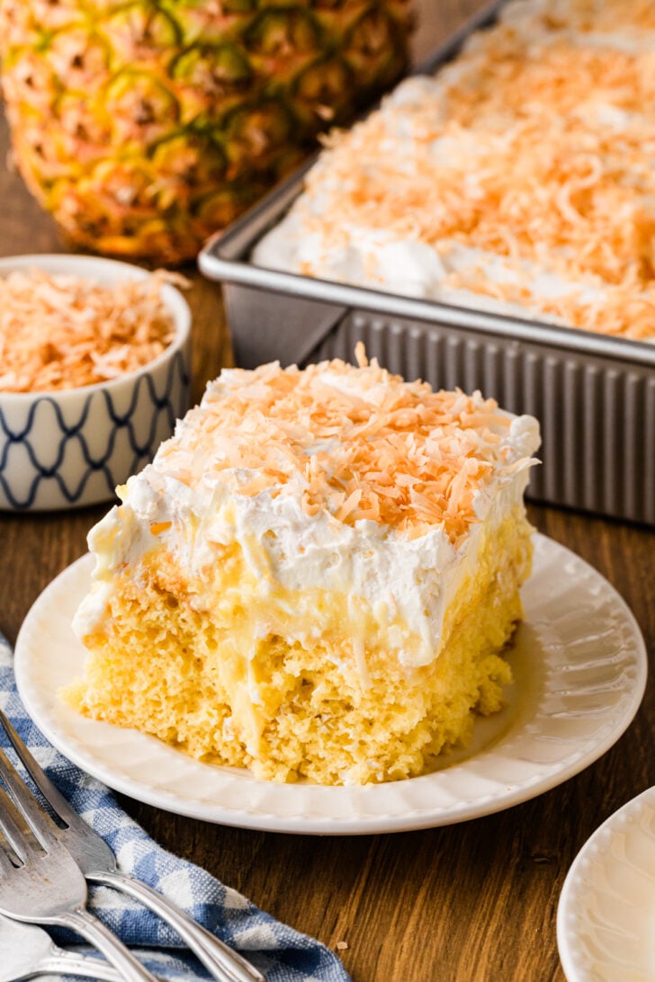 Pineapple coconut poke cake topped with whipped cream and toasted coconut flakes in a 13x9 pan and white plate.