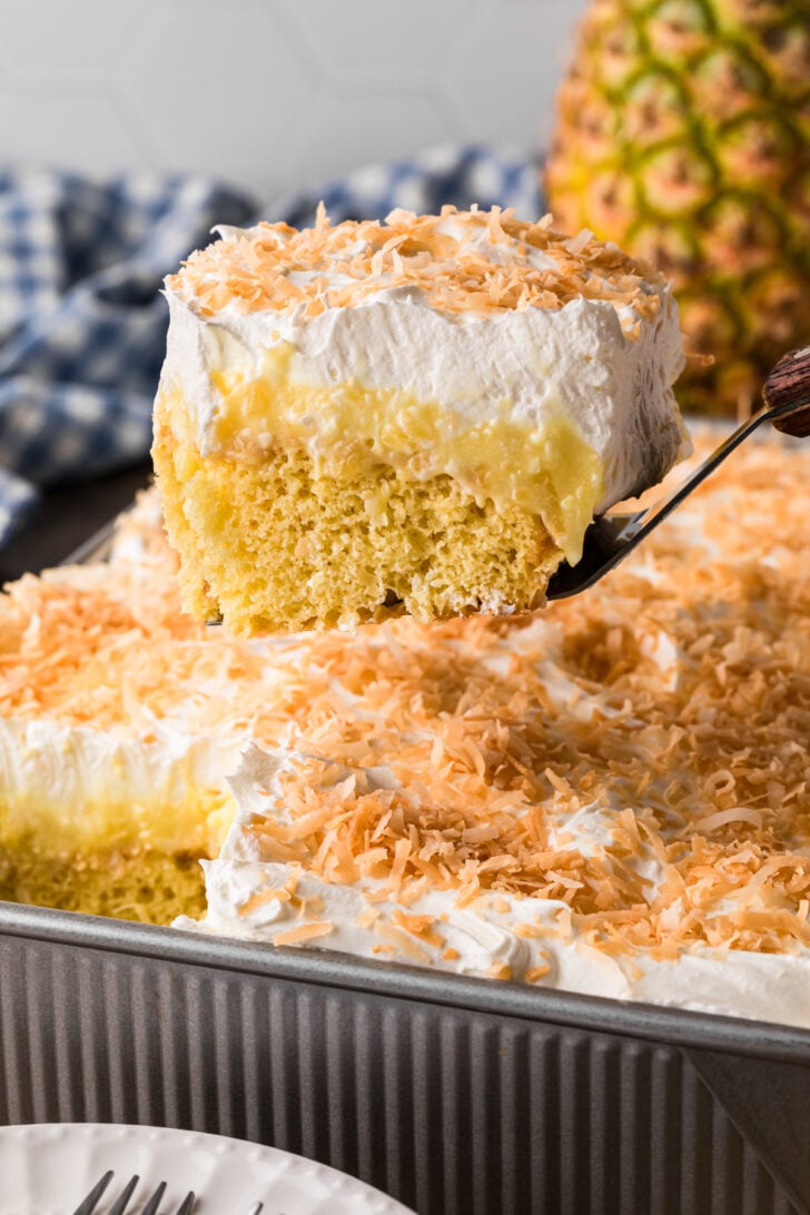 Slice of pineapple coconut poke cake on a cake server showing creamy coconut pudding layers and toasted coconut topping.