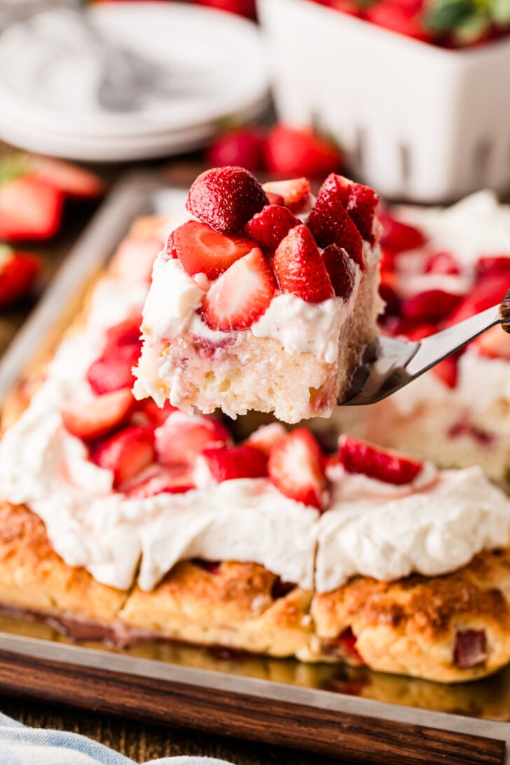 Fork lifting slice of Strawberry Shortcake Sheet Cake topped with whipped cream and fresh berries from wooden serving board