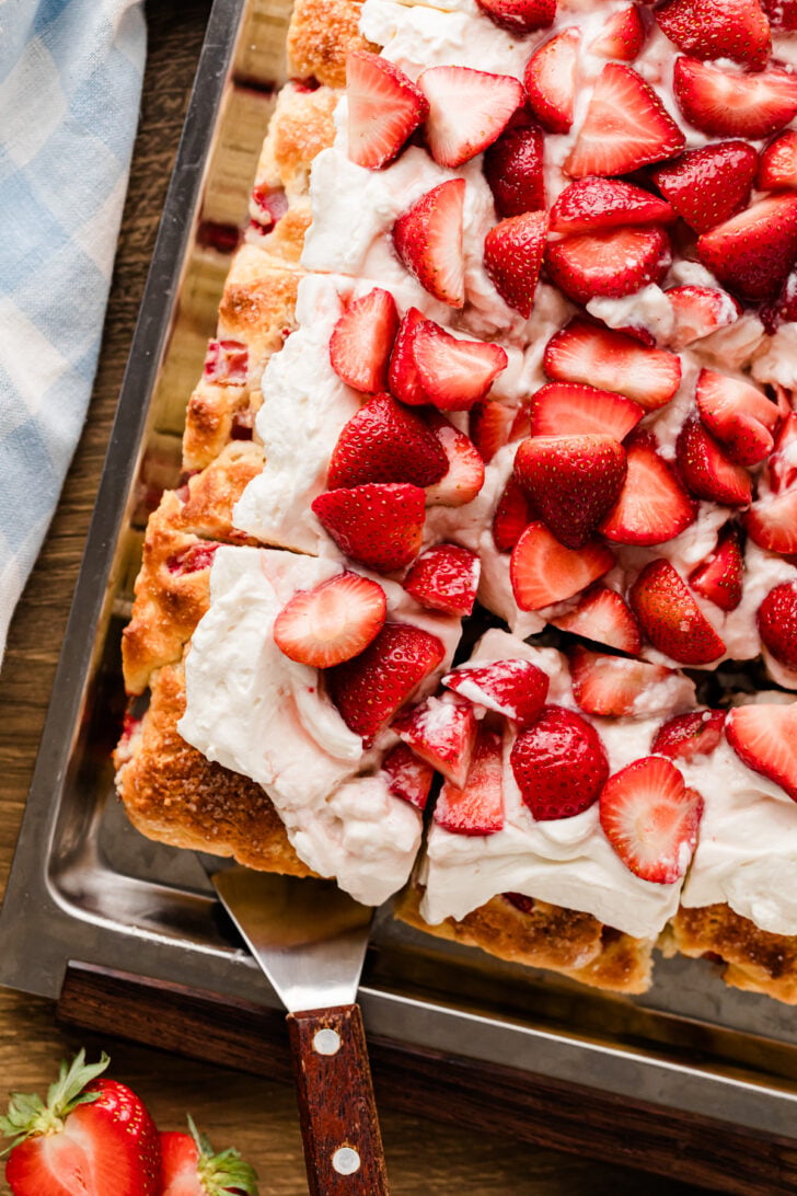 Strawberry Shortcake Sheet Cake with whipped cream topping and fresh sliced strawberries on a metal baking sheet with a spatula