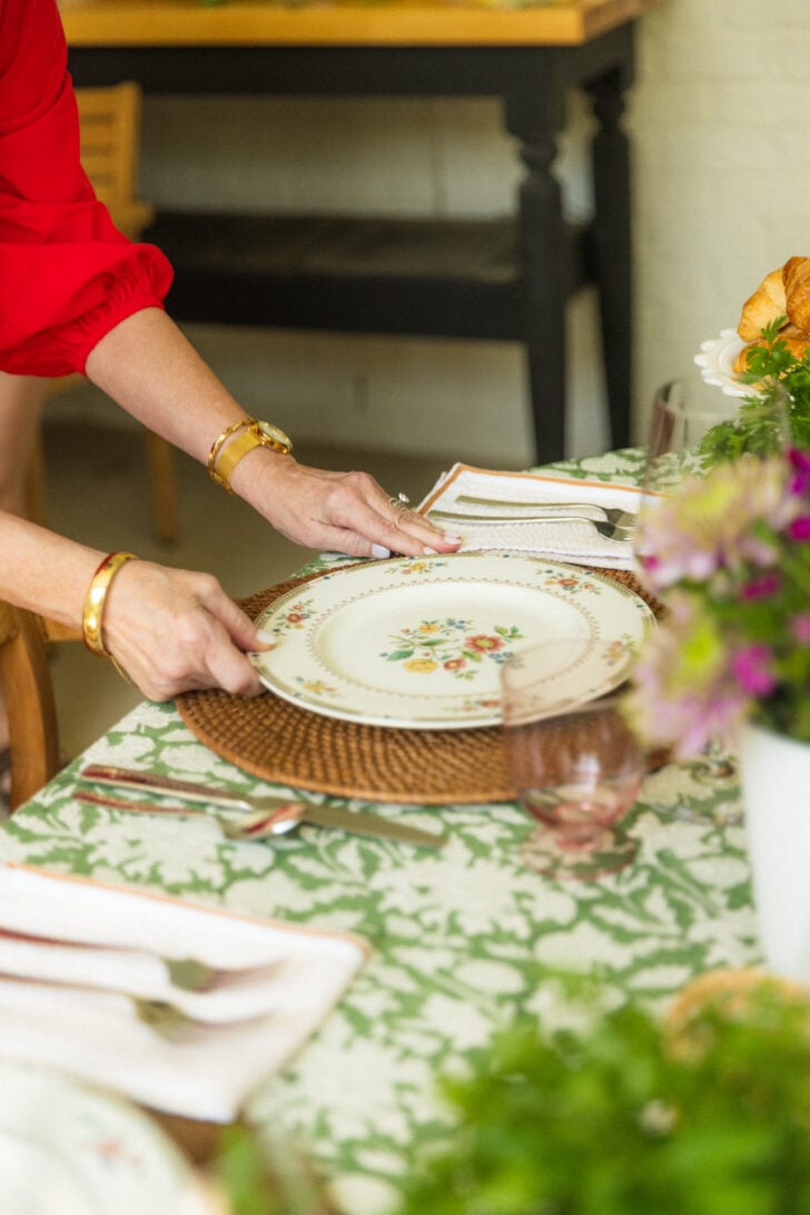Hands setting a floral plate on a green tablecloth with woven placemats and fresh flowers.