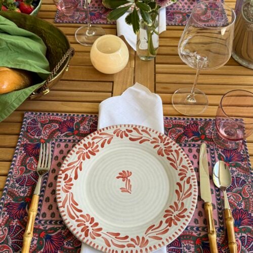 Elegant outdoor table setting with a floral placemat, patterned plate, gold flatware, and candlelight.