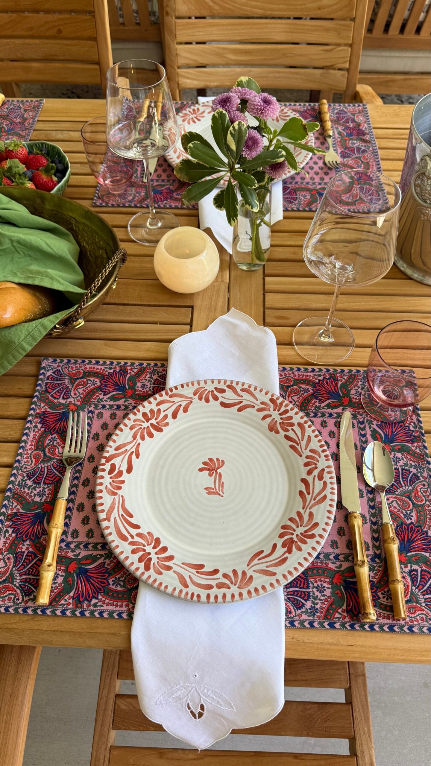 Elegant outdoor table setting with a floral placemat, patterned plate, gold flatware, and candlelight.