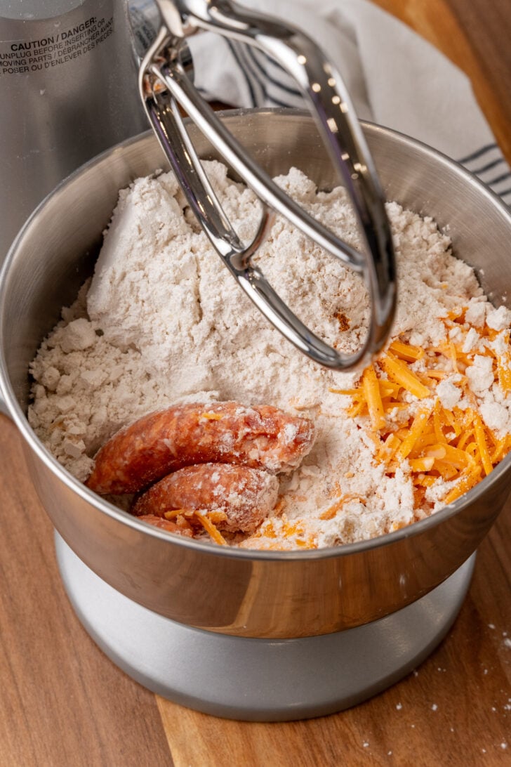 Overhead view of cheesy sausage balls 4 ingredients in metal mixing bowl: flour, shredded cheddar cheese, and raw sausage with electric mixed.