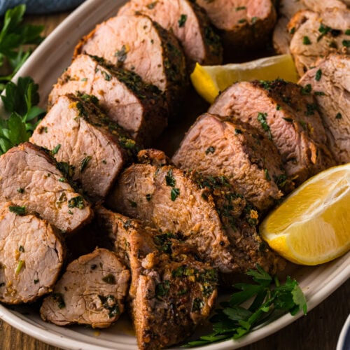 Sliced roasted pork tenderloin on a platter with lemon wedges and fresh parsley.