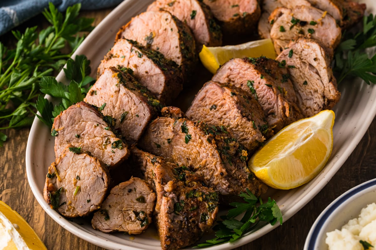 Sliced roasted pork tenderloin on a platter with lemon wedges and fresh parsley.