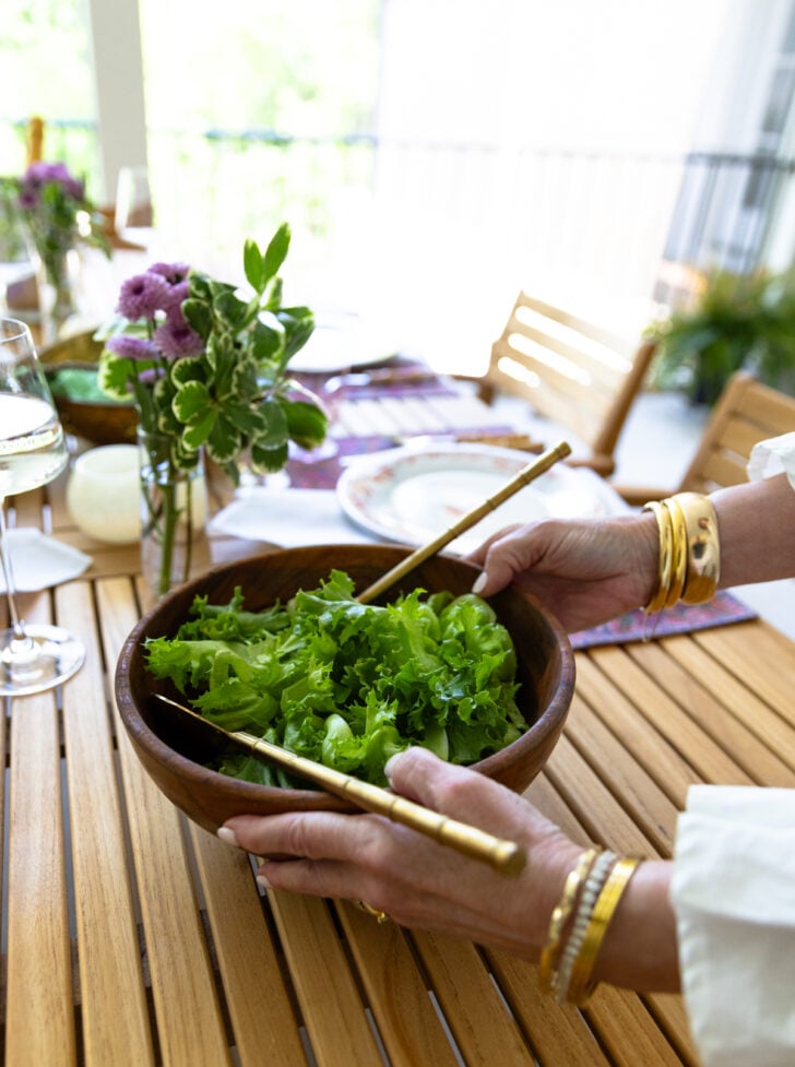 Hands tossing a green salad in a wooden bowl at an outdoor spring table setting.