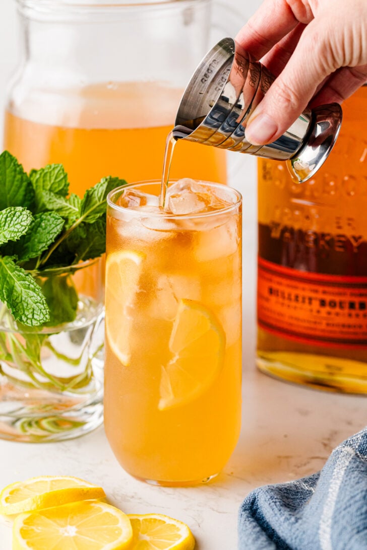 Pouring bourbon into an Arnold Palmer cocktail with lemon and mint.