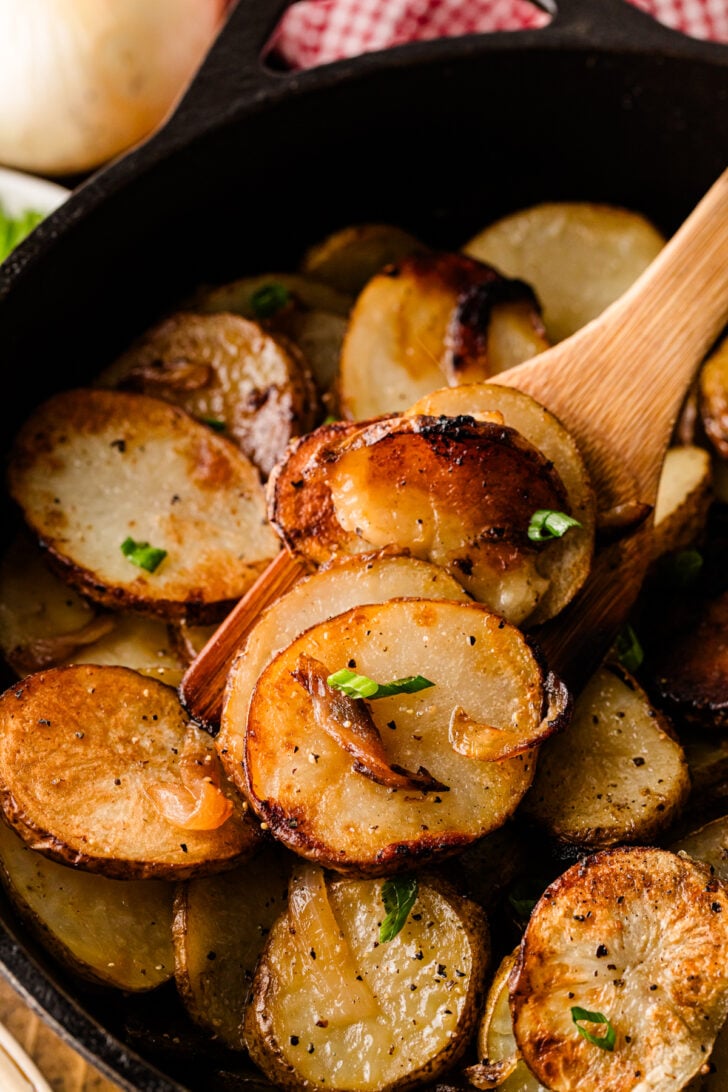 Wooden spoon lifting crispy fried potatoes and onions from a cast iron skillet