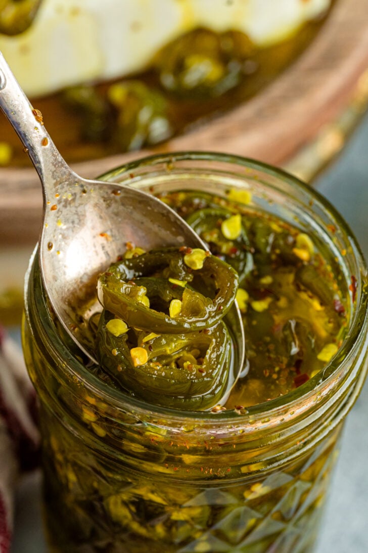 Jar of candied jalapeños beside cream cheese topped with cowboy candy, fresh jalapeños, and crackers on a green serving plate.